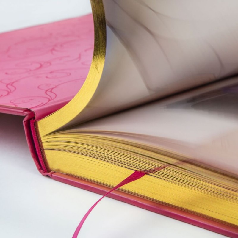 Nahaufnahme eines geöffneten Buchs mit goldenen Seitenrändern, rosa Einband und rosa Lesebändchen.